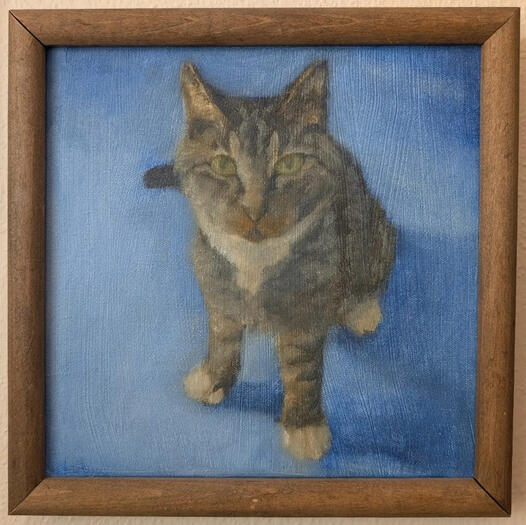 photo of a framed oil painting on a beige apartment wall; ~2013, oils on canvas depicting my old cat, Xena. a perspective view, she is sitting on a blue rug, looking up at the viewer with her big green eyes. she has black stripes over her grayish brown fur